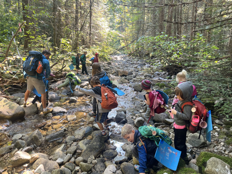Children crossing a rocky creek together during outdoor learning at Roots To Sky Forest School near Nelson BC